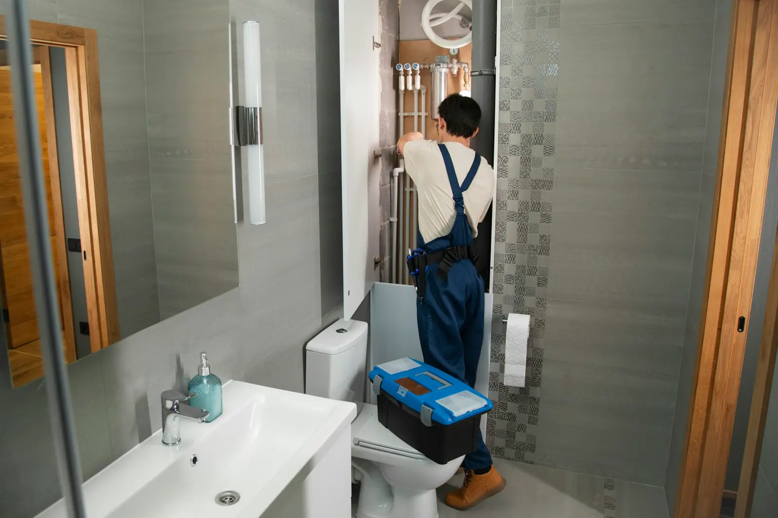 Plumber in overalls with a toolbox repairing plumbing in a small bathroom