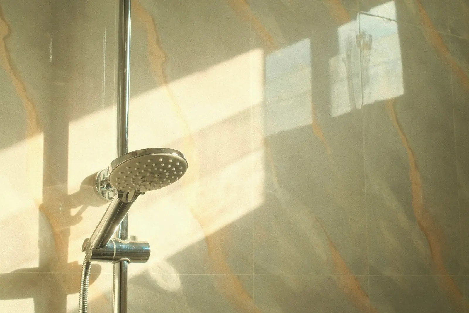 Shower handset on a riser rail lit by sunlight on a marble wall